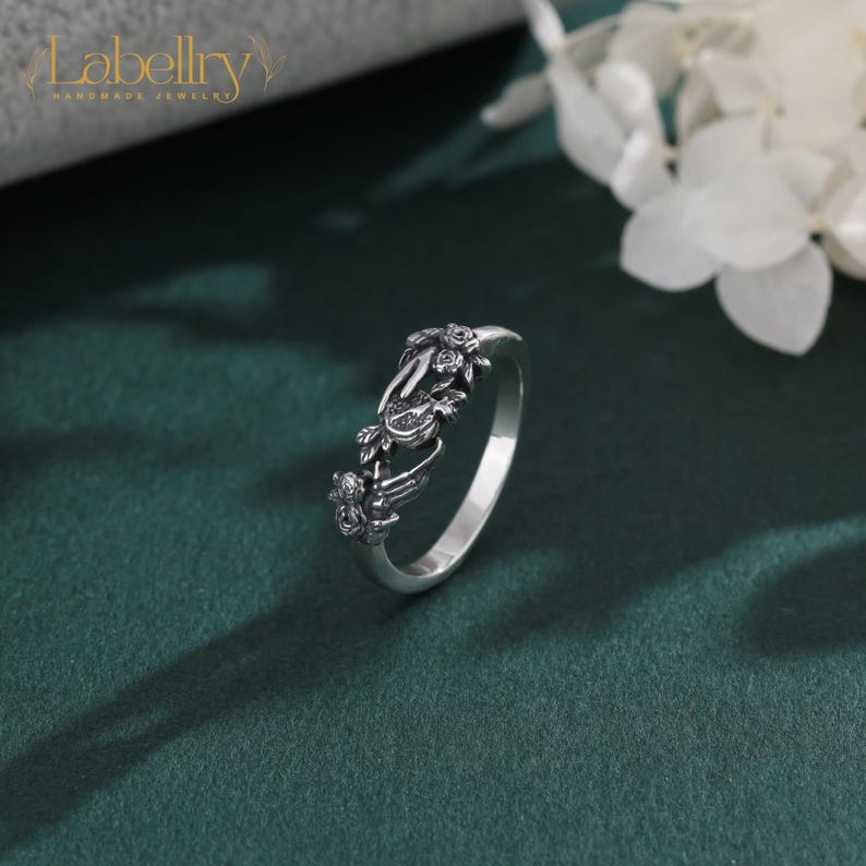 May include: A silver ring featuring a detailed, oxidized metal design of two hands reaching toward each other, accented with small sculpted rose motifs. The band has a polished finish on the interior and a dark, textured finish on the exterior.