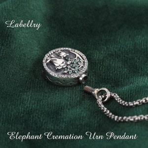 May include: A silver-toned Elephant Cremation Urn Pendant with a detailed floral design. The pendant is round with a screw-top closure and is attached to a chain. The word "Labellry" is in the upper left corner.