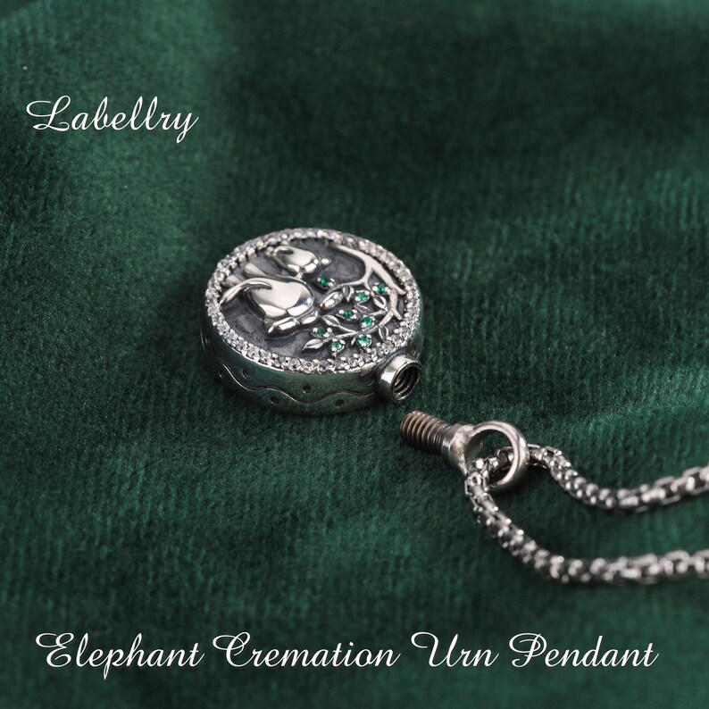 May include: A silver-toned Elephant Cremation Urn Pendant with a detailed floral design. The pendant is round with a screw-top closure and is attached to a chain. The word "Labellry" is in the upper left corner.