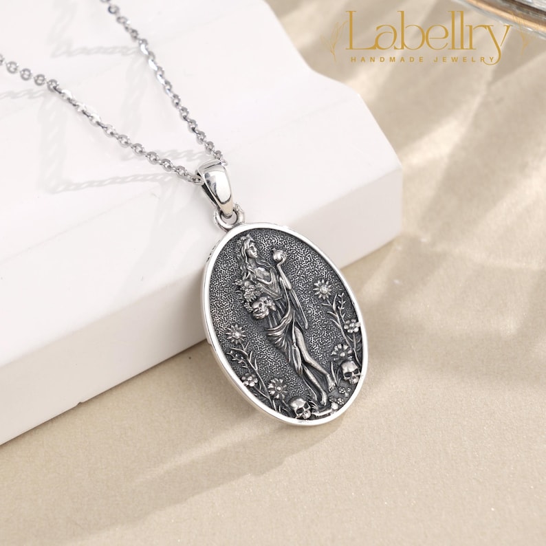 May include: Silver-toned pendant necklace featuring an oval pendant with a detailed relief of a woman holding flowers and a skull. The pendant is surrounded by floral and skull motifs. The chain is delicate and silver.