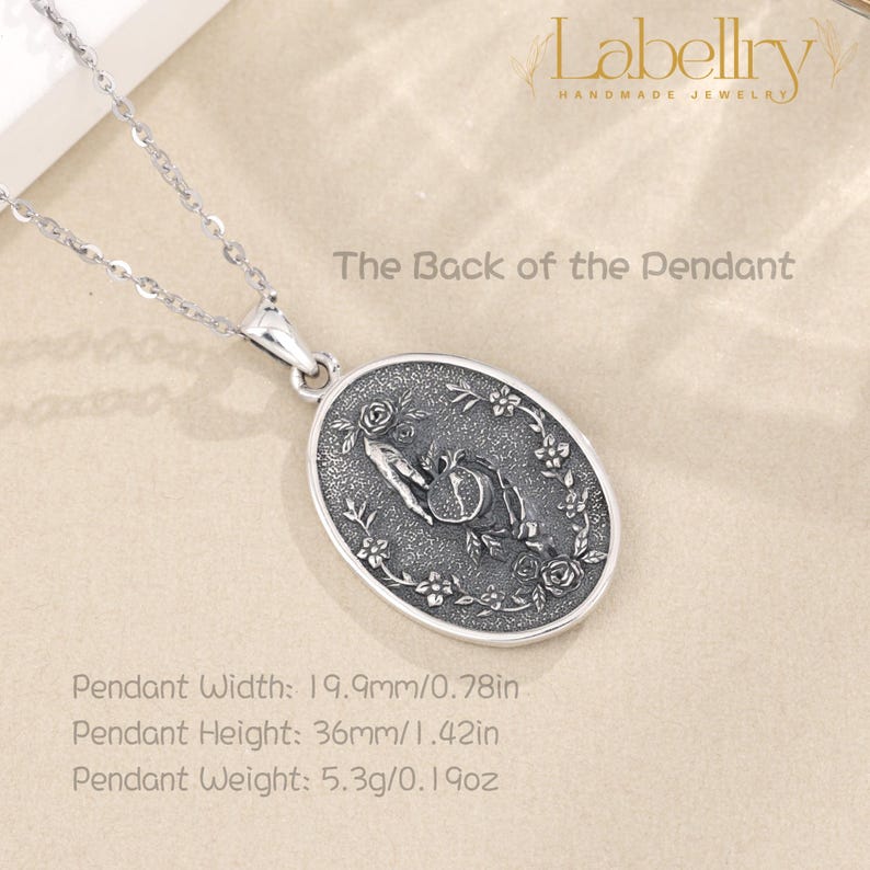 May include: Silver pendant necklace with an oval pendant featuring a hand holding a pomegranate, surrounded by floral details. The pendant is 0.78 inches wide and 1.42 inches high. The text "The Back of the Pendant" is visible.