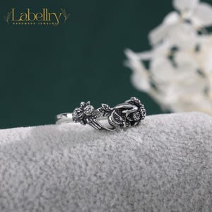 May include: An adjustable silver-toned ring featuring a detailed design of two hands holding a cluster of roses. The metal has an oxidized finish to highlight the intricate textures of the floral elements and the fingers. The word Adjustable is printed.