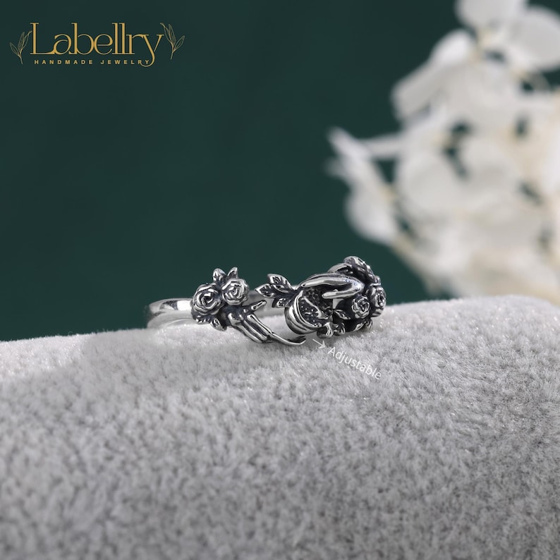 May include: An adjustable silver-toned ring featuring a detailed design of two hands holding a cluster of roses. The metal has an oxidized finish to highlight the intricate textures of the floral elements and the fingers. The word Adjustable is printed.
