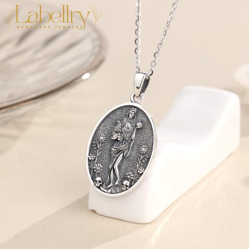 May include: Silver pendant necklace with an oval pendant featuring a detailed relief of a woman holding flowers, surrounded by floral and skull motifs. The necklace has a delicate chain and the brand name "Labellry" is visible.