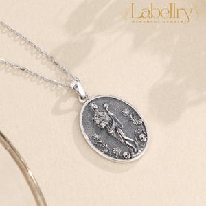 May include: A silver pendant necklace featuring an oval pendant with a detailed relief design. The pendant depicts a figure holding flowers, surrounded by floral elements and skulls. The necklace has a delicate chain and the brand name "Labellry Handmade Jewelry" is visible.