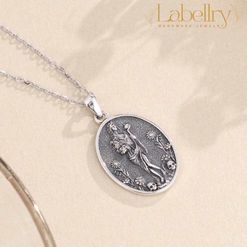 May include: A silver pendant necklace featuring an oval pendant with a detailed relief design. The pendant depicts a figure holding flowers, surrounded by floral elements and skulls. The necklace has a delicate chain and the brand name "Labellry Handmade Jewelry" is visible.