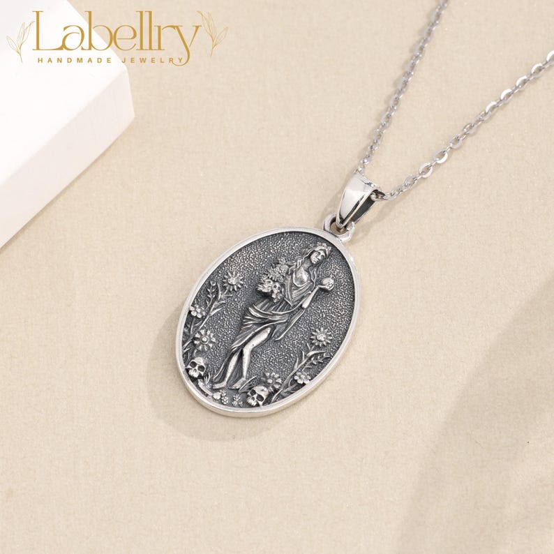 May include: A silver-toned pendant necklace featuring an oval pendant with a detailed relief of a woman holding flowers, surrounded by floral and skull motifs. The necklace has a delicate chain and the brand name "Labellry" is visible in the upper left corner.