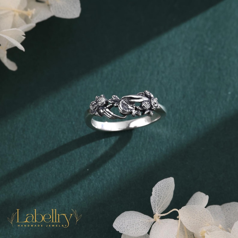 May include: A silver-toned metal ring featuring a detailed design of two hands reaching toward each other, surrounded by small sculpted rose motifs. The band has an oxidized finish that highlights the intricate textures of the floral and hand elements.