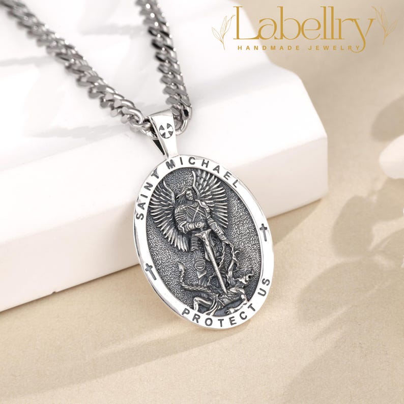 Pu&ograve; includere: Collana color argento con un ciondolo ovale raffigurante San Michele. Il ciondolo reca la scritta "SAINT MICHAEL PROTECT US". La catena &egrave; a maglia grossa. Gioielli del marchio Labellry.