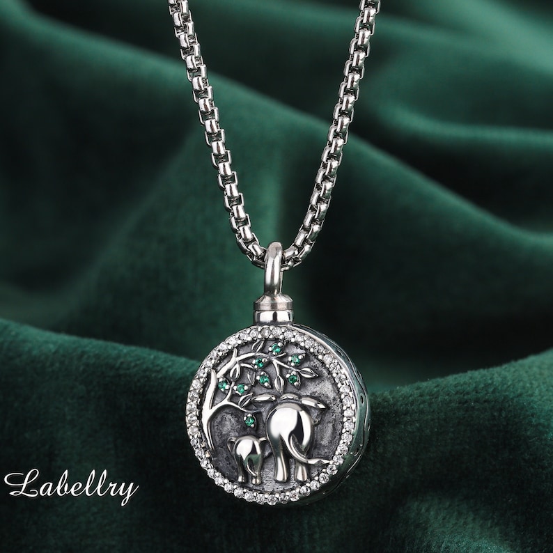 May include: Silver-toned pendant necklace featuring a round charm with an elephant and baby elephant design under a tree with green accents. The charm is surrounded by small, clear stones. The necklace has a chain link style.