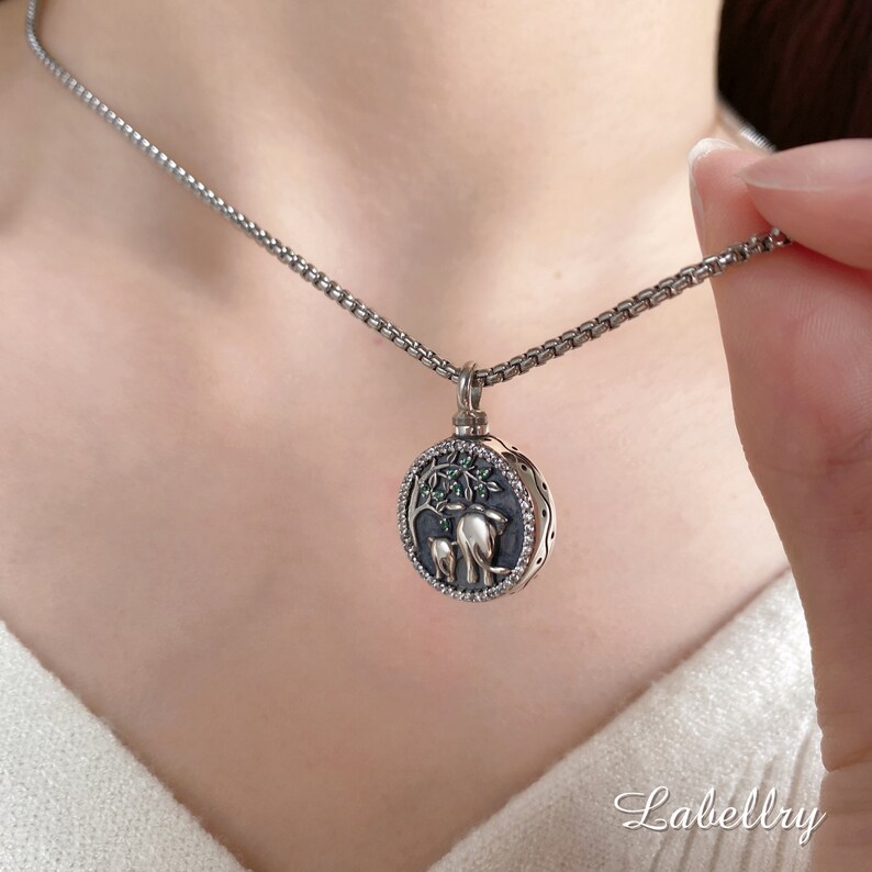 May include: A silver-toned necklace with a round pendant featuring an elephant and calf under a tree. The pendant is detailed with small green and clear stones around the edge and hangs from a chain.