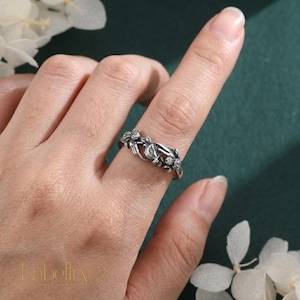 May include: A silver-toned metal ring featuring a detailed, sculpted design of two hands meeting in the center, accented with small floral motifs. The band has an oxidized finish that highlights the intricate textures and shapes of the metalwork.