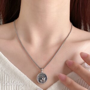 May include: Silver-toned necklace with a round pendant featuring a tree and two animals. The pendant is encircled by small, clear stones. The necklace is worn on a person.