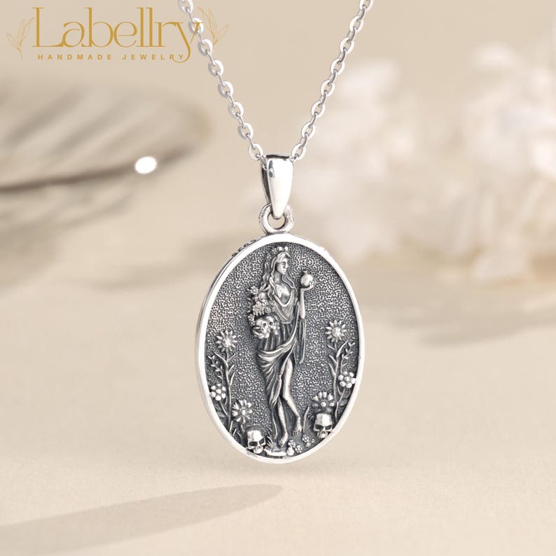 May include: A silver-toned pendant necklace featuring an oval pendant with a detailed relief of a woman holding flowers and a sphere. The pendant is surrounded by floral and skull motifs, with the brand name "Labellry" at the top.