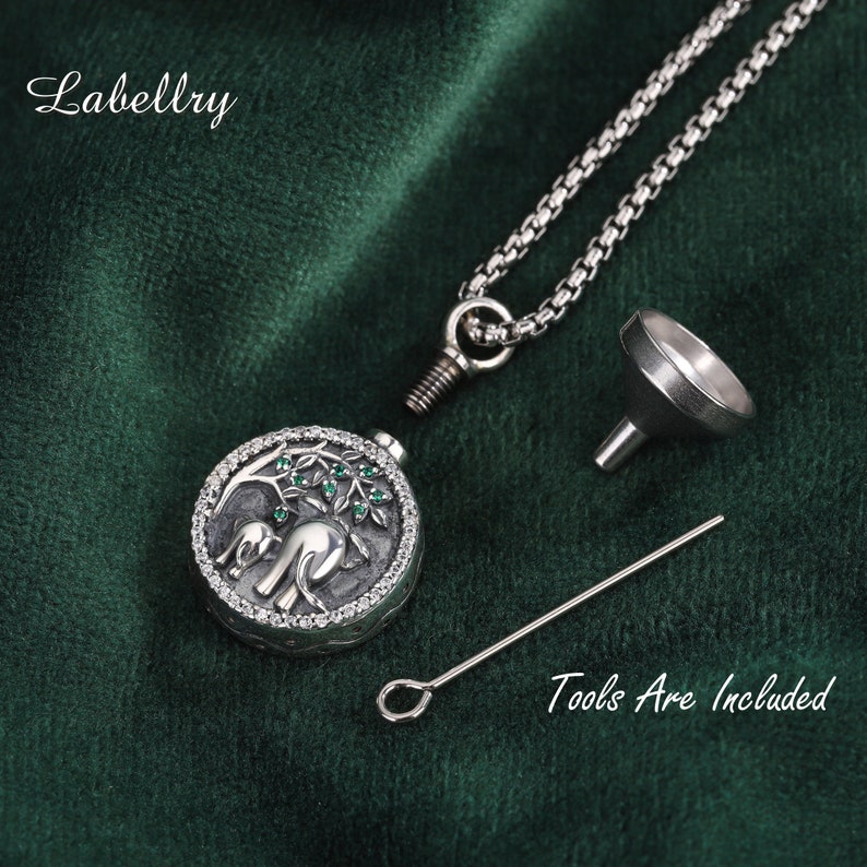 May include: A silver-colored pendant necklace with a round locket featuring an elephant design with green accents. The necklace includes a chain, a small funnel, and a tool. The text "Tools Are Included" is visible.