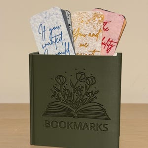 Op de afbeelding: Een olijfgroene rechthoekige houder met het woord BOOKMARKS en een gegraveerde illustratie van een open boek met bloemen die uit de pagina's komen. Verschillende rechthoekige bladwijzers met diverse kleuren en tekst zitten in de houder.