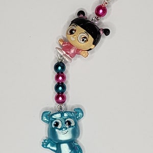 Puede incluir: Un charm con los personajes Boo y Sulley de la película de animación Monstruos, S.A. Boo lleva un vestido rosa y Sulley es de color azul claro. El charm está conectado con cuentas rosas y turquesas y un cierre plateado.
