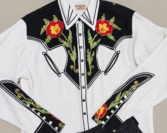 May include: A long-sleeved white shirt with black shoulder panels and cuffs, featuring red and yellow floral embroidery with green leaves. The shirt has a button-down front with black buttons and a label reading Sam & Sons Co. Black pinstriped trousers.