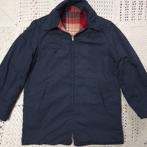 Op de afbeelding: Marineblauwe jas met een volledige ritssluiting en een kraag. De jas heeft een rood, bruin en zwart geruite voering. Het heeft twee voorzakken en een borstzak. De jas is van gemiddeld gewicht en lijkt gemaakt van een duurzaam materiaal.