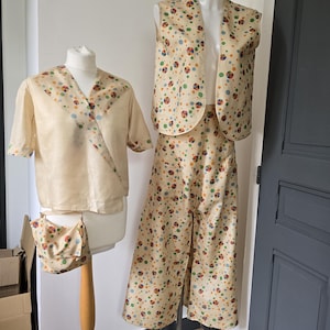 Op de afbeelding: Een vintage crèmekleurige outfit met een kleurrijk stippenpatroon. De set bevat een wikkel top met korte mouwen, een vest en een broek met wijde pijpen. Een kleine bijpassende tas wordt ook getoond. De outfit wordt op mannequins gepresenteerd, wat de retrostijl laat zien.