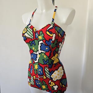 Op de afbeelding: Een vintage badpak uit één stuk met een levendig, abstract patroon. Het badpak heeft een rode basis met kleurrijke vormen in groen, blauw, geel en wit. Het heeft een halternek en een getailleerd silhouet, weergegeven op een mannequin.