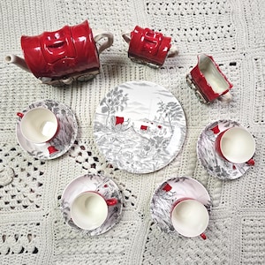 May include: A red ceramic tea set with a whimsical design, including a teapot, creamer, sugar bowl, and teacups with saucers. The plates and saucers feature a black and white pastoral scene. The set is arranged on a white crocheted blanket.