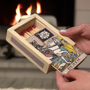 Peut inclure: Une boîte d'allumettes en bois avec un grattoir à allumettes rayé rouge et blanc. La boîte d'allumettes est ouverte et une carte de tarot avec l'image de la carte de la Mort est tirée. La carte est une carte de tarot de style Rider-Waite traditionnel avec un chevalier noir monté sur un cheval blanc. La carte est intitulée "DEATH."
