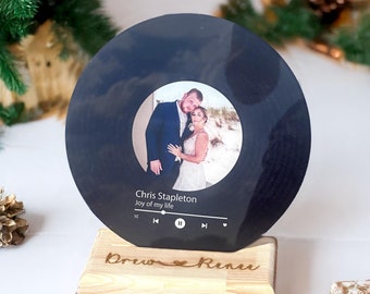 May include: A black vinyl record with a photo of a couple in front of a beach. The record is on a wooden stand with the names "Drew" and "Renee" engraved on it. The record also has a music player interface with the text "Chris Stapleton Joy of my life".