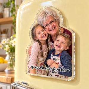 May include: A custom-shaped acrylic photo magnet featuring a smiling grandmother with a young girl and boy wearing aprons. The image displays the text Grandma's Sunshine in a black script font at the bottom, attached to a yellow surface.