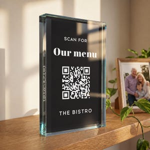Op de afbeelding: Een rechthoekig, helder acrylblok met een zwarte inzet met witte tekst die luidt SCAN FOR Our menu, een centrale QR-code en de tekst THE BISTRO onderaan. Het bord heeft een dik, transparant frame met afgeschuinde randen.