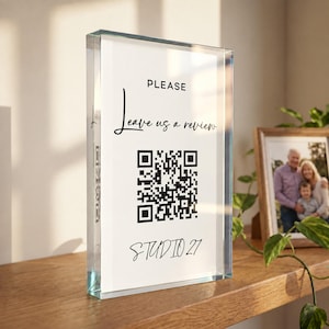 Könnte beinhalten: Ein rechteckiges Schild aus klarem Acryl mit einem weißen Einleger, der den schwarzen Text PLEASE Leave us a review, einen zentralen schwarz-weißen QR-Code und den Text STUDIO 27 am unteren Rand aufweist, aufrecht stehend.