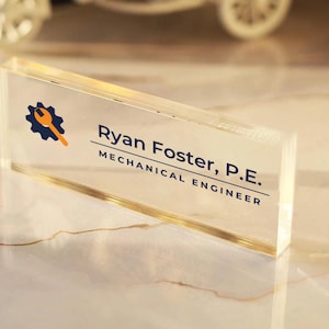 May include: A rectangular clear acrylic desk nameplate featuring a blue gear icon with an orange wrench. The text reads Ryan Foster, P.E. and MECHANICAL ENGINEER in dark blue lettering, centered on the transparent block surface.