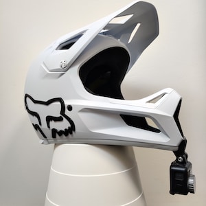 Könnte beinhalten: Weißer Integral-Motorradhelm mit schwarzem Fox-Logo und einer schwarzen Action-Kamera, die an der Seite montiert ist.