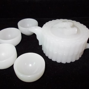 May include: A white ceramic tea set featuring a teapot with a lid and four small, rounded cups. The teapot has a textured, ribbed design and a curved handle. The set is displayed on a dark surface.