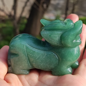 Op de afbeelding: Een gebeeldhouwd groen edelsteenbeeldje van een mythisch wezen, mogelijk een draak of leeuw. Het beeldje wordt in een hand gehouden, waarbij de gedetailleerde kenmerken en het gepolijste oppervlak worden benadrukt. De edelsteen heeft een doorschijnende kwaliteit en de gravure is gedetailleerd.