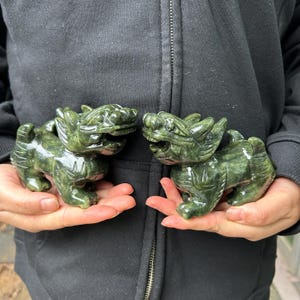 Op de afbeelding: Twee gebeeldhouwde jade beeldjes van mythische wezens, elk met een gedetailleerde draakachtige kop en een stevig lichaam. De beeldjes zijn diepgroen van kleur en lijken in iemands handen te worden gehouden. De gravures hebben ingewikkelde details en een gepolijste afwerking.