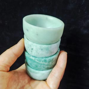 Op de afbeelding: Vier kleine, ronde kommen in jadegroen, op elkaar gestapeld. De kommen worden in een hand gehouden en hebben elk een iets andere tint groen, variërend van licht tot donker. De kommen hebben een gladde, gepolijste afwerking.