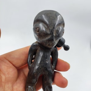 May include: A dark grey figurine of an alien-like figure with large eyes and a small, rounded head. The figure has a small, rounded appendage extending from its mouth. The figurine is held in a hand against a white background.