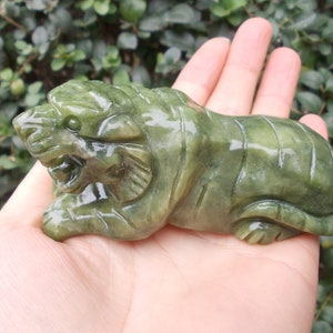 Pode incluir: Uma estatueta de tigre de jade esculpida em tons de verde. O tigre é retratado em uma pose reclinada com traços faciais e marcas corporais detalhadas. A escultura é segurada em uma mão, mostrando seu tamanho e artesanato.
