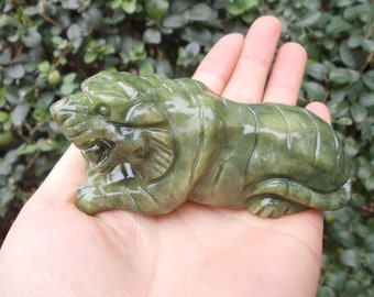 May include: A carved jade tiger figurine in shades of green. The tiger is depicted in a reclining pose with detailed facial features and body markings. The sculpture is held in a person's hand, showcasing its size and craftsmanship.
