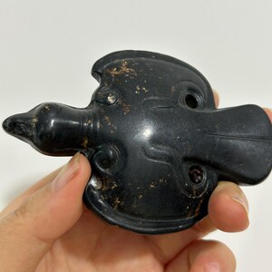 May include: A black, duck-shaped carving with a smooth, polished surface. The carving has a detailed beak, head, and body, with small holes and intricate designs. The object is held in a hand against a white background.