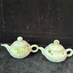 May include: Two small, light green teapots with lids and rounded handles. The teapots have a speckled pattern of darker green and are made of a stone-like material. The spouts are curved and the lids have small, round knobs.