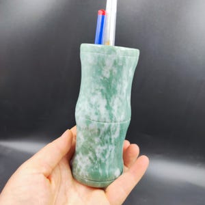 Puede incluir: Un portalápices de piedra moteada verde y blanca con un diseño similar al bambú. El portalápices es cilíndrico con una forma ligeramente curvada y contiene varios bolígrafos. El portalápices está hecho de piedra maciza.