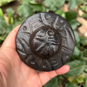 Könnte beinhalten: Dunkles, kreisförmiges Objekt mit geprägtem Design. Es hat einen erhöhten Mittelbereich mit radialen Linien und kleinen Punkten am Umfang. Die Farbe ist dunkel, möglicherweise metallisch. Gehalten in einer Hand.