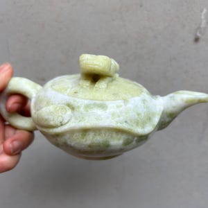 Puede incluir: Una pequeña tetera tallada intrincadamente en piedra verde pálido. La tetera presenta un pico curvo, un cuerpo redondeado y un asa de bucle. Un pequeño elemento decorativo se encuentra sobre la tapa. La tetera es sostenida por una mano.