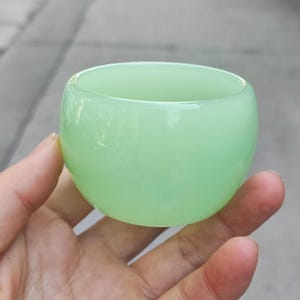 May include: A small, rounded, light green glass cup. The cup has a smooth, glossy surface and a wide opening. The cup is held in a hand, with the background out of focus.