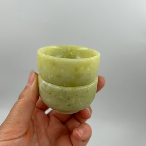 May include: Two small, round, jade-colored bowls stacked on top of each other. The bowls have a smooth, polished finish and a light green hue with subtle variations in color. The bowls are held in a hand against a plain white background.