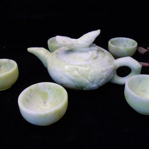 Puede incluir: Un juego de té verde claro con una tetera y cuatro tazas pequeñas. La tetera tiene una tapa con forma de pájaro y tallas florales. Las tazas y la tetera están hechas de una piedra moteada de color verde claro. El juego se exhibe sobre una superficie negra.