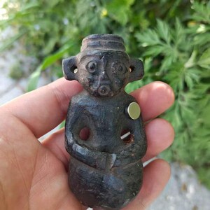 May include: A small, dark-colored figurine of a seated figure with a hat and large ears. The figurine has a hole through the body and is held in a hand against a blurred green background. A small, round, silver-colored magnet is attached to the figurine.
