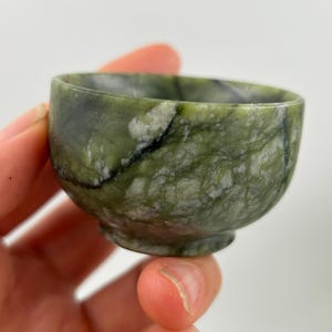 Op de afbeelding: Een kleine, ronde groene stenen kom met een voet. De kom heeft een gemarmerd uiterlijk met tinten groen, wit en zwart. De kom wordt in een hand gehouden.